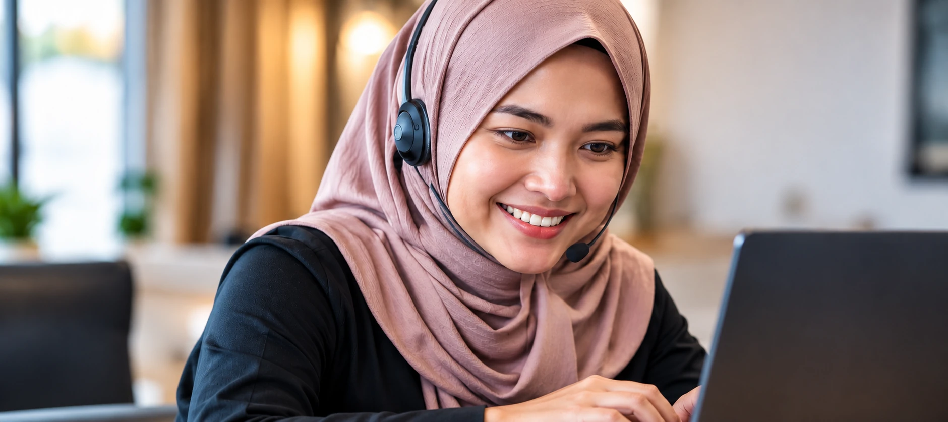 Wanita Malaysia memakai headset sambil mengurus pertanyaan pelanggan dan WhatsApp di komputer riba